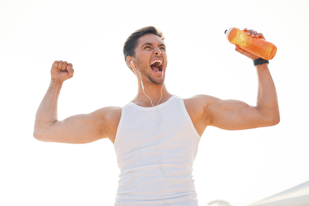 Lekkoatletycznego mięśni mężczyzna uczucie zwycięstwa i cieszy się sukces trzyma butelkę wody. Emocji, energii, gest - młody człowiek świętuje zwycięstwo i krzyczy z słońca światła w tle. - Zdjęcie, obraz