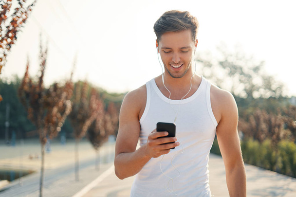 Shot of an attractive, smiling, sporty young man starting his music play list on the smartphone while looking to screen outdoors. - Photo, Image