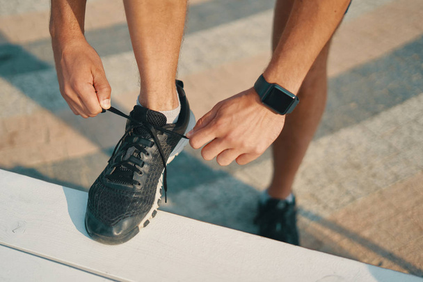 Sportsman ties his black sneakers. Athletic man tying shoelaces while getting ready to run. Smart watch on left hand. Closeup shot - 写真・画像