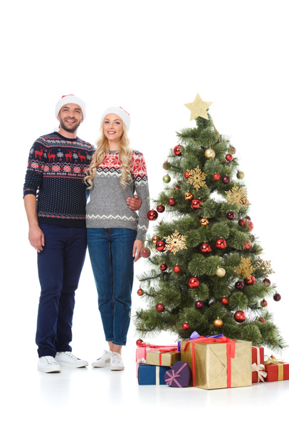 beautiful happy couple in santa hats standing near christmas tree with gifts, isolated on white - Photo, Image