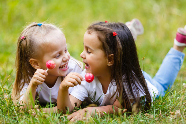 Two little girls relaxing and eating a lollipops at a park - Photo, Image