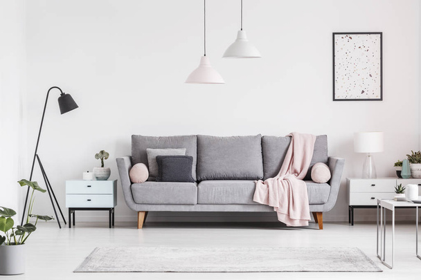 Grey couch with blanket between cabinets in living room interior with poster and lamps. Real photo - Zdjęcie, obraz