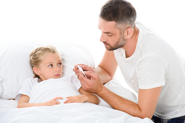 father looking at electronic thermometer while adorable ill daughter with temperature lying in bed, isolated on white - Photo, Image