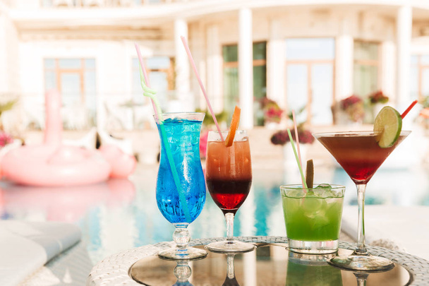 Photo of four cocktail drinks in glasses at table near hotel swimming pool during sunny summer day - Фото, изображение