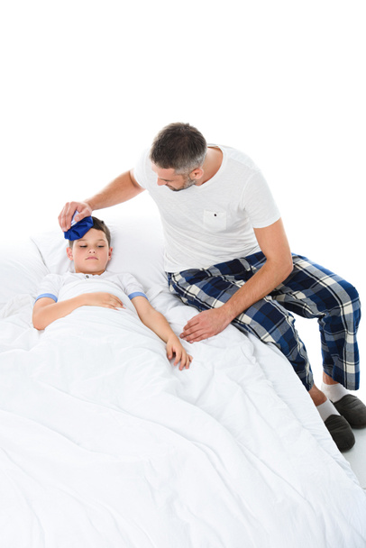 father holding ice pack on head of sick son with temperature lying in bed, isolated on white - Фото, изображение
