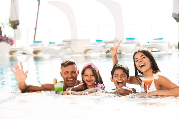 Happy young family having fun inside a swimming pool outdoors in summer with cocktails, waving hands - Фото, зображення