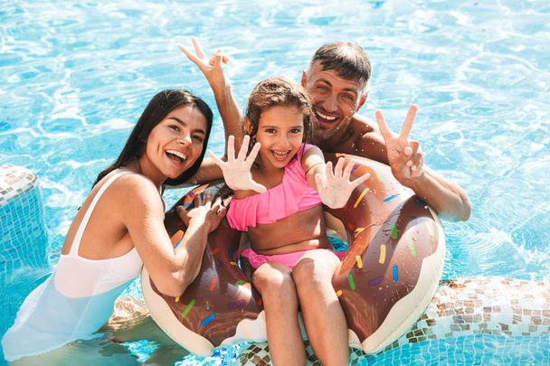 Cheerful young family having fun together at the swimming pool outdoors in summer, swimming with inflatable ring donut, waving hands - Photo, Image