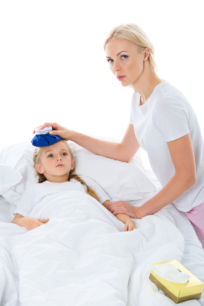 madre sosteniendo la bolsa de hielo en la cabeza de la hija enferma con la temperatura acostada en la cama, aislada en blanco
 - Foto, Imagen