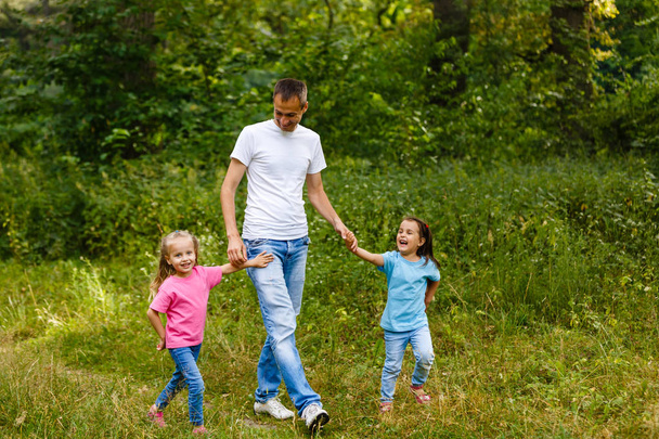 Père heureux tenant la main de deux petites filles marchant dans un parc verdoyant
 - Photo, image
