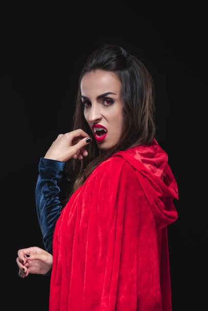 dreadful vampire woman in red cloak showing her teeth isolated on black - Photo, Image
