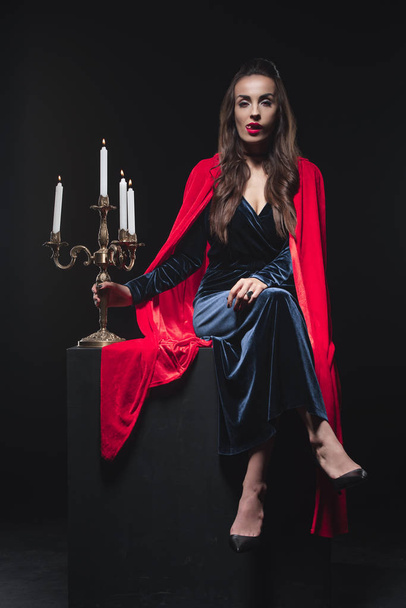 Hermoso vampiro de capa roja lamiendo dientes y sosteniendo candelabro antiguo aislado en negro
 - Foto, Imagen