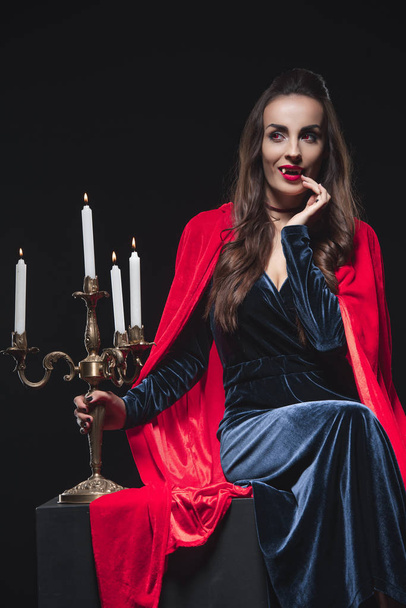 vampire in red cloak holding candelabrum and touching her fangs isolated on black - Photo, Image