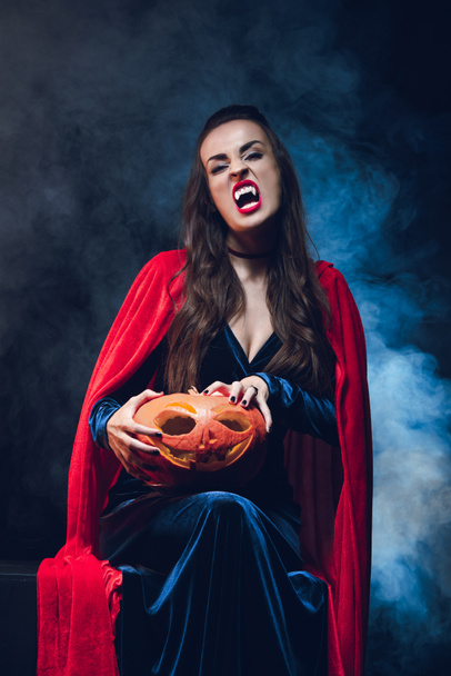 woman in vampire costume holding jack o lantern on darkness with smoke  - Фото, изображение