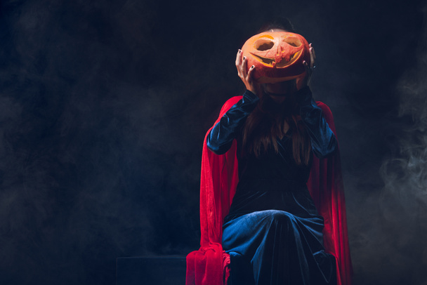 mujer en capa roja sosteniendo jack o linterna en frente de la cara en la oscuridad con humo
  - Foto, imagen
