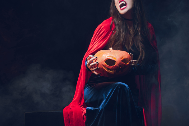 cropped view of beautiful woman in vampire costume holding pumpkin on darkness with smoke  - Foto, afbeelding
