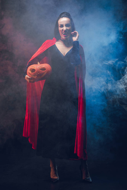 atractiva mujer en traje de vampiro celebración de calabaza sobre fondo oscuro con humo
  - Foto, imagen