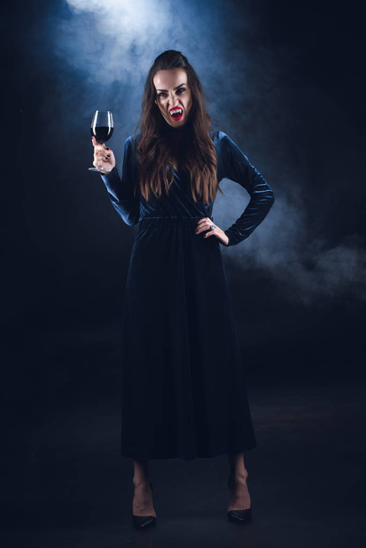 ζοφερή βαμπίρ κρατώντας wineglass με αίμα σε σκούρο φόντο με καπνό  - Φωτογραφία, εικόνα