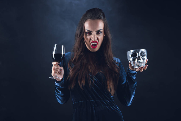 Vampir hält Metallschädel und Weinglas mit Blut in der Dunkelheit mit Rauch  - Foto, Bild