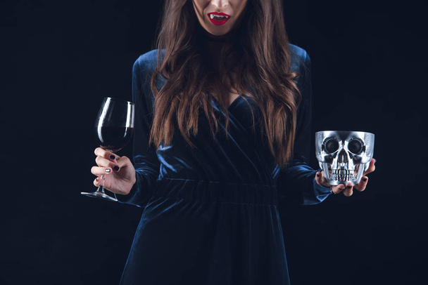 bijgesneden weergave van vampier vrouw houdt van schedel en wijnglas met bloed geïsoleerd op zwart - Foto, afbeelding
