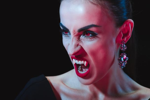 dreadful vampire showing her fangs and looking away isolated on black - Foto, imagen