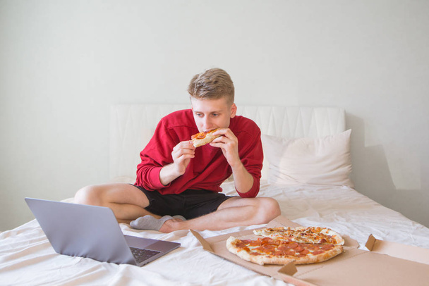 Привлекательный молодой человек сидит на кровати в уютной светлой комнате, сосредоточен на просмотре фильма на ноутбуке и поедании пиццы из коробки. Выходные с фильмами и коробкой пиццы. Доставка пиццы
 - Фото, изображение