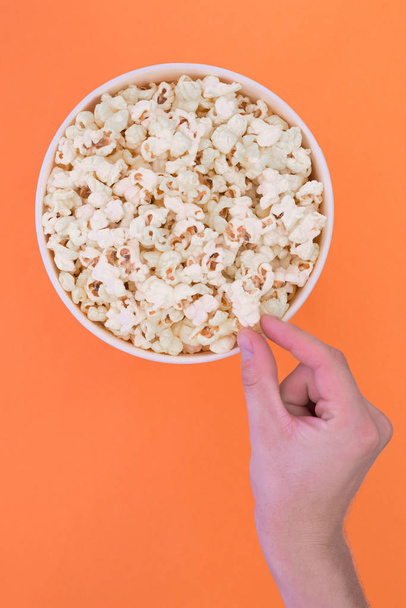 Male hand takes a popcorn from a paper cup on a orange background. Man eats popcorn. Isolated. Cinema Concept. Flat lay. Copyspace. - Fotografie, Obrázek