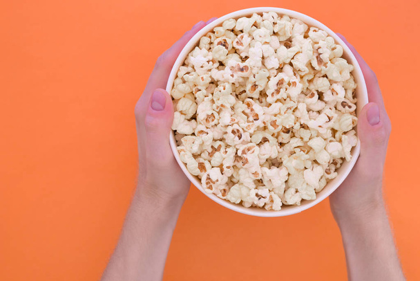 Men's hands hold a paper cup with popcorn on an orange background. Man with popcorn in his hands.solated. Cinema Concept. Flat lay. Copyspace. - 写真・画像