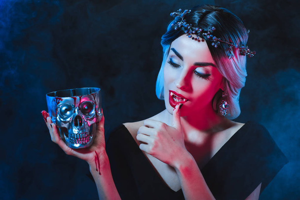 beautiful vampire woman holding metal skull with blood and licking her fingers isolated on black - Fotó, kép