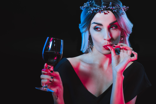 Vampirfrau hält Weinglas mit Blut und leckt ihre Finger isoliert auf schwarz - Foto, Bild