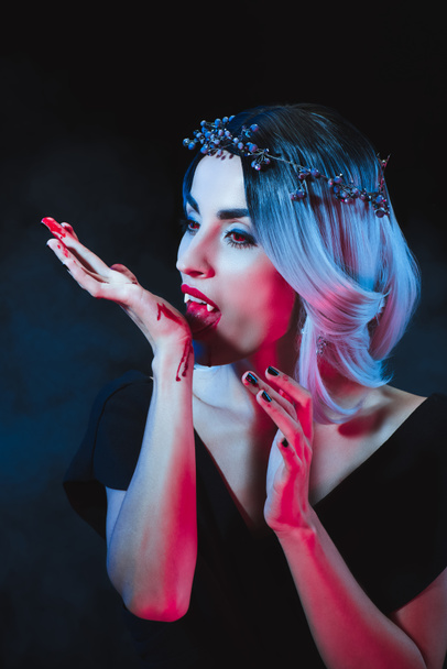 attractive vampire licking blood from hand on dark background with smoke  - Фото, изображение