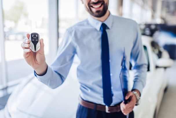 recortado disparo de vendedor sonriente sosteniendo las llaves del coche y portapapeles en salón de concesionarios
 - Foto, Imagen