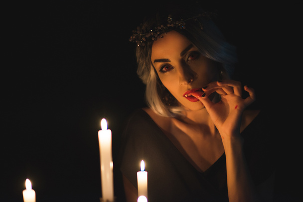 beautiful woman in vampire costume licking her fingers with candles on foreground isolated on black - Fotografie, Obrázek
