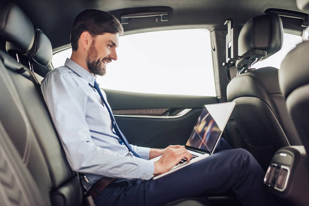 Widok z boku z młody biznesmen Brodaty działa na laptopie w samochodzie - Zdjęcie, obraz