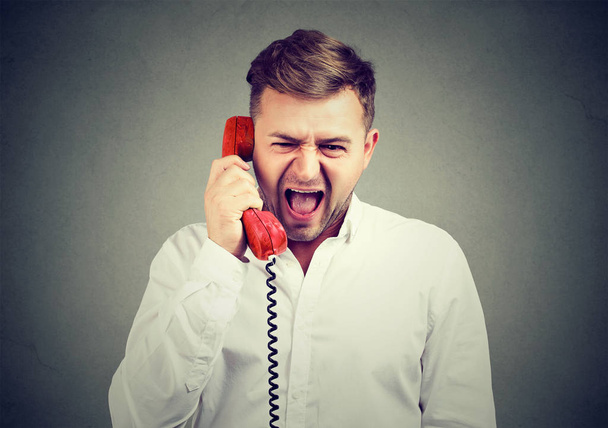 Young man in white shirt holding red telephone handset and screaming in frustration on gray background.  - 写真・画像