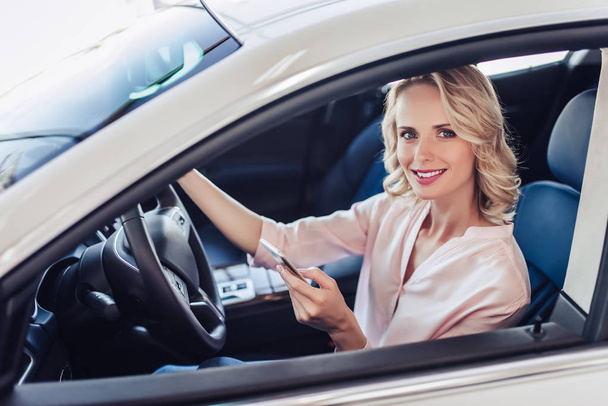 portrait of attractive caucasian woman sitting in the car, using smartphone and lookin at the camera - Foto, Imagem