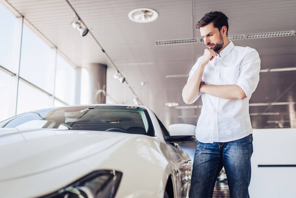 portrait of pensive male client looking at car in dealership salon - Фото, изображение