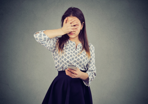 Young woman making mistake while using smartphone and looking sad on gray background - Photo, image