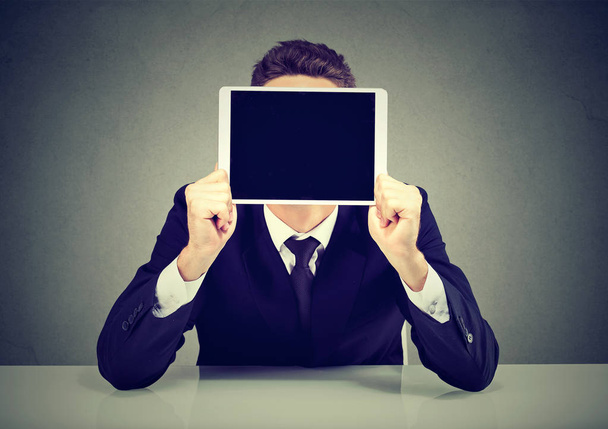 Anonymous businessman in suit holding tablet in front of face sitting on gray background - Photo, image
