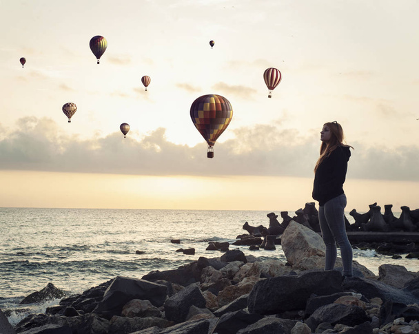 Mujer mirando a globos de aire caliente volando sobre el océano al amanecer
. - Foto, imagen