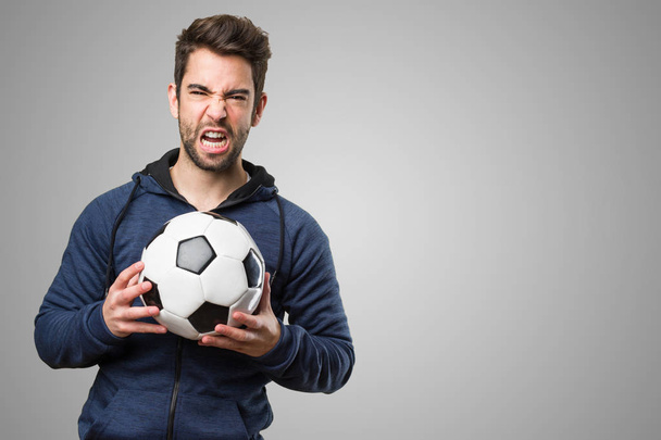 θυμωμένος νεαρός κρατώντας μια μπάλα ποδοσφαίρου σε γκρίζο φόντο - Φωτογραφία, εικόνα