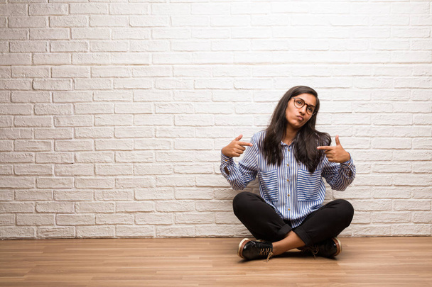 Mujer india joven sentarse contra una pared de ladrillo orgulloso y seguro, señalando los dedos, ejemplo a seguir, concepto de satisfacción, arrogancia y salud
 - Foto, imagen