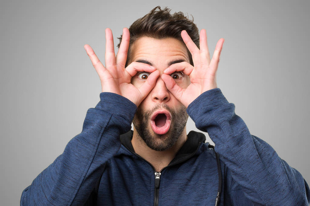 verrast jonge man doen bril gebaar op grijze achtergrond - Foto, afbeelding