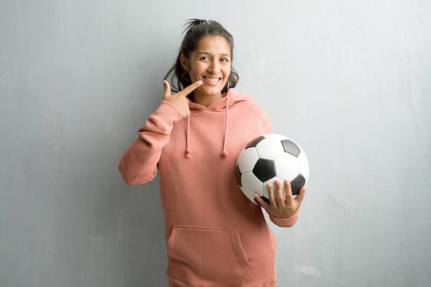 Mladý sportovní indiánka proti zdi úsměvy, ukazující ústa, koncept dokonalých zubů, bílé zuby, má veselá a žoviální přístup. Držení míče Fotbal hrát hru. - Fotografie, Obrázek