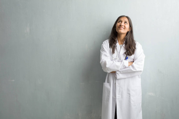 Jeune femme médecin indienne contre un mur levant les yeux, pensant à quelque chose d'amusant et ayant une idée, concept d'imagination, heureux et excité
 - Photo, image