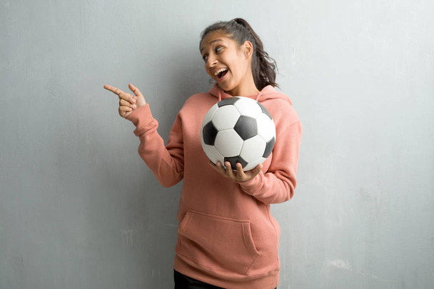 Молодая спортивная индианка прислонилась к стене, указывая в сторону, улыбаясь удивленно представляя что-то, естественное и случайное. Проведение футбольный мяч, чтобы играть в игру
. - Фото, изображение