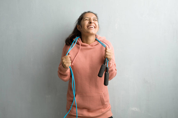 Nuori urheilullinen intialainen nainen seinää vasten erittäin onnellinen ja innoissaan, nostaa aseita, juhlii voittoa tai menestystä, voittaa lotossa. Hyppynarua pidellen
. - Valokuva, kuva