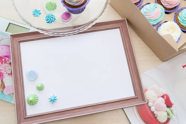 wooden frame with white sheet around meringue and cupcake with multi-colored cream, kitchen scale, magazine - Photo, Image