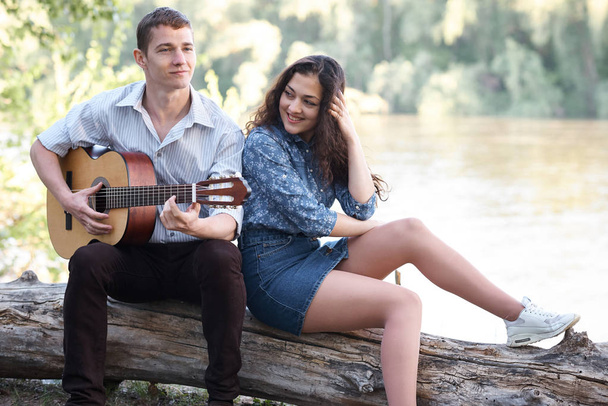 junges Paar, das auf einem Baumstamm am Fluss sitzt und Gitarre spielt, sommerliche Natur, helles Sonnenlicht, Schatten und grüne Blätter, romantische Gefühle - Foto, Bild