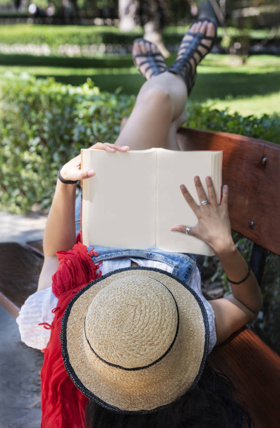 όμορφη νεαρή γυναίκα με ένα ψάθινο καπέλο διαβάζοντας ένα βιβλίο σε ένα παγκάκι, στο πάρκο - Φωτογραφία, εικόνα