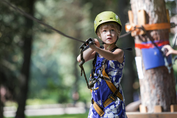 Young child girl in safety harness and helmet attached to cable moves carefully along rope way in recreation park on green bokeh background. Sport, game and leisure concept - Фото, изображение
